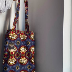 Pode incluir: Uma bolsa tote azul e vermelha com um padr&atilde;o geom&eacute;trico. A bolsa tem uma etiqueta branca com o texto "Bijou Crafted" nela.