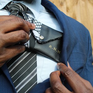 May include: A man wearing a blue suit jacket with a white shirt and a black and white striped tie. He is holding a black leather wallet with the initials "R.H.H." embossed in gold.