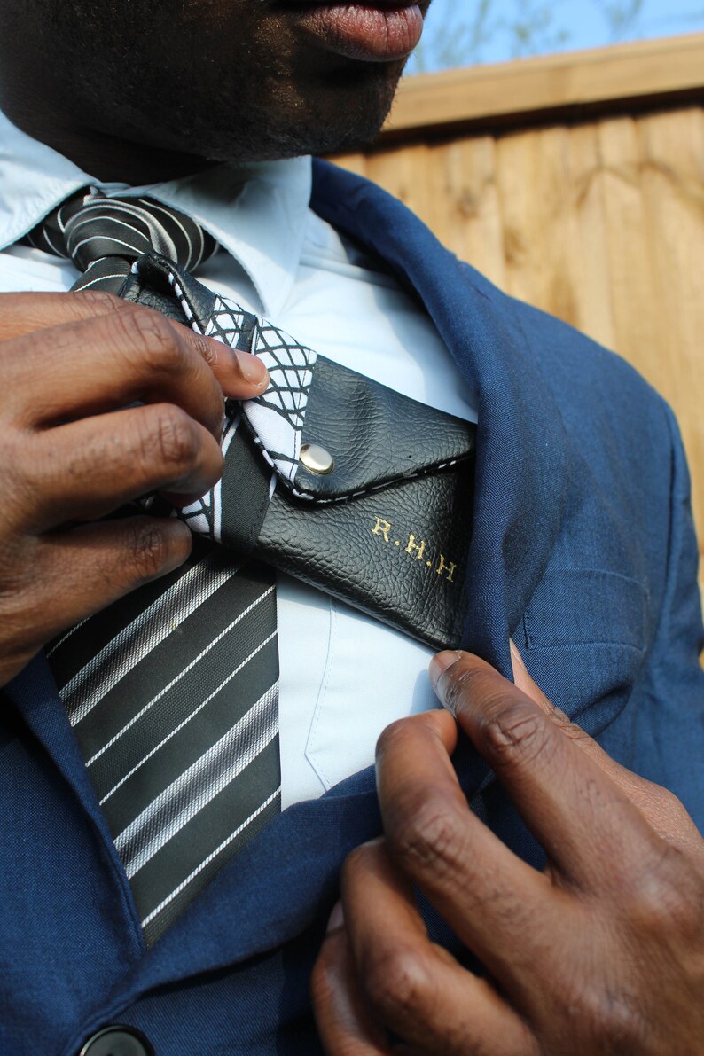May include: A man wearing a blue suit jacket with a white shirt and a black and white striped tie. He is holding a black leather wallet with the initials "R.H.H." embossed in gold.
