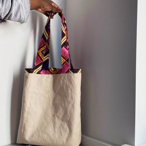 Pode incluir: Uma bolsa tote de lona bege com al&ccedil;as de tecido estampado em rosa, amarelo, azul e preto.