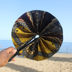 May include: A round hand fan with a black handle and a patterned fabric. The fabric features a dark brown background with yellow and black geometric designs. The fan is held up against a blue sky and ocean background.