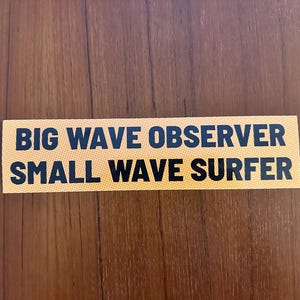 May include: A rectangular sticker with the text "BIG WAVE OBSERVER" above "SMALL WAVE SURFER" in black, on a yellow background. The sticker is placed on a brown wooden surface.