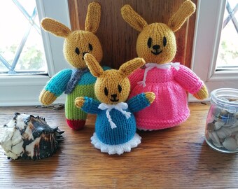 May include: Three knitted bunny rabbits, one in blue, one in pink, and one in green and yellow. The bunnies are sitting on a wooden surface.