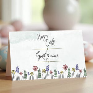 May include: White card with the text "Happy Easter" and "Guest's name" with a line of colorful flowers at the bottom.