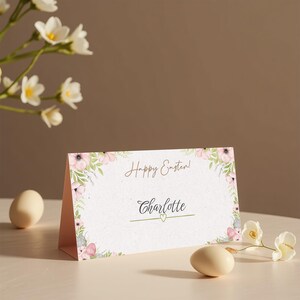 May include: A pink and green floral design place card with the text "Happy Easter!" and "Charlotte" in a gold font. The place card is sitting on a light brown surface.