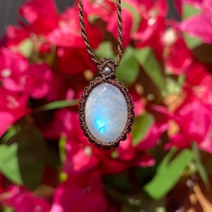 May include: An oval moonstone pendant featuring a blue iridescent sheen, encased in a dark brown macrame cord setting. The pendant hangs from a matching twisted brown cord necklace, with a small silver-toned bead accent above the stone.