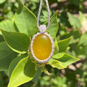 May include: A yellow oval gemstone pendant with a macrame knot design, hanging from a gray cord.