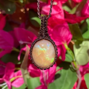 May include: A pendant featuring an oval-shaped, iridescent opal gemstone with flashes of green and orange, secured in a dark brown macrame cord setting. The stone is suspended from a matching brown braided cord necklace.