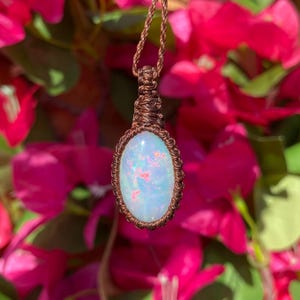 May include: A handcrafted pendant necklace featuring an oval opal gemstone. The opal is set in a macrame weave with a brown cord. The opal has a light blue base color with pink and orange flecks. The background is a blur of pink flowers.