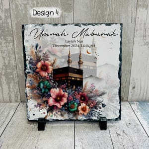 Pu&ograve; includere: Una targa in ardesia bianca con un motivo floreale e un'immagine della Kaaba a La Mecca. Il testo "Umrah Mubarak" &egrave; scritto in calligrafia nella parte superiore della targa. Il testo "Laylah Nur Dicembre 2024/1446 AH" &egrave; scritto sotto l'immagine.