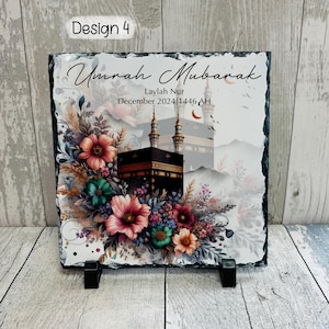 May include: A black slate plaque with a white background featuring a floral design and a depiction of the Kaaba in Mecca. The text "Umrah Mubarak" is written in a decorative font, along with the name "Laylah Nur" and the date "December 2024/1446 AH".