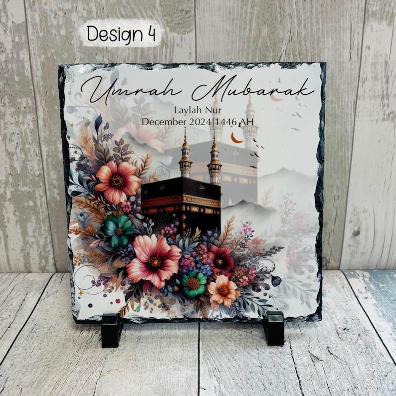 May include: A black slate plaque with a white background featuring a floral design and a depiction of the Kaaba in Mecca. The text "Umrah Mubarak" is written in a decorative font, along with the name "Laylah Nur" and the date "December 2024/1446 AH".