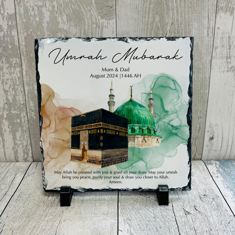 May include: A black slate plaque with a green and white watercolour background featuring a mosque and the Kaaba. The text "Umrah Mubarak" is written in cursive at the top, followed by "Mum & Dad" and "August 2024 | 1446 AH". The bottom of the plaque reads "May Allah be pleased with you & grant all your duas. May your umrah bring you peace, purify your soul & draw you closer to Allah. Ameen."