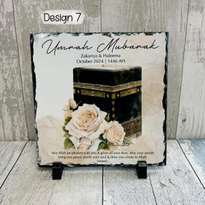 Pu&ograve; includere: Una targa in ardesia bianca con un'immagine nera e dorata della Kaaba, un motivo floreale bianco e il testo "Umrah Mubarak Zakariya & Haleema Ottobre 2024 | 1446 AH" e "Che Allah sia contento di te e ti conceda tutte le tue suppliche. Che la tua Umrah ti porti pace, purifichi la tua anima e ti avvicini ad Allah. Amen."