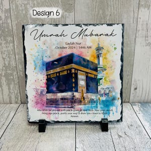 May include: A black slate plaque with a watercolor painting of the Kaaba in Mecca. The plaque has the text "Umrah Mubarak" and "Laylah Nur October 2024 | 1446 AH" and a message wishing the person a blessed Umrah.