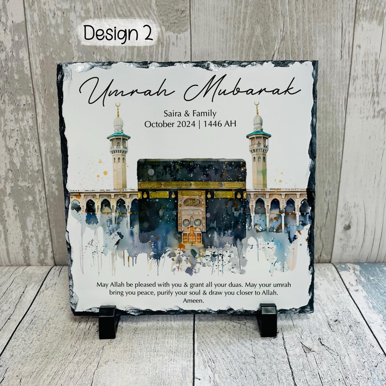 Pu&ograve; includere: Una targa in ardesia nera con un'illustrazione ad acquerello della Kaaba a La Mecca, in Arabia Saudita. Il testo "Umrah Mubarak" &egrave; scritto in un carattere decorativo nella parte superiore della targa. Il testo "Saira & Family October 2024 | 1446 AH" &egrave; scritto sotto l'illustrazione. La targa include anche un messaggio di benedizioni e una preghiera per la pace e la purificazione.