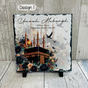 Pu&ograve; includere: Una placca in ardesia nera con un'immagine colorata della Kaaba a La Mecca, circondata da fiori e uccelli. Il testo "Umrah Mubarak" &egrave; scritto in uno stile calligrafico decorativo, seguito da "Talib & Nazia" e "Dicembre 2024 | 1446 AH".