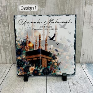 May include: A black slate plaque with a colorful image of the Kaaba in Mecca, surrounded by flowers and birds. The text "Umrah Mubarak" is written in a decorative font, along with the names "Talib & Nazia" and the date "December 2024 | 1446 AH".
