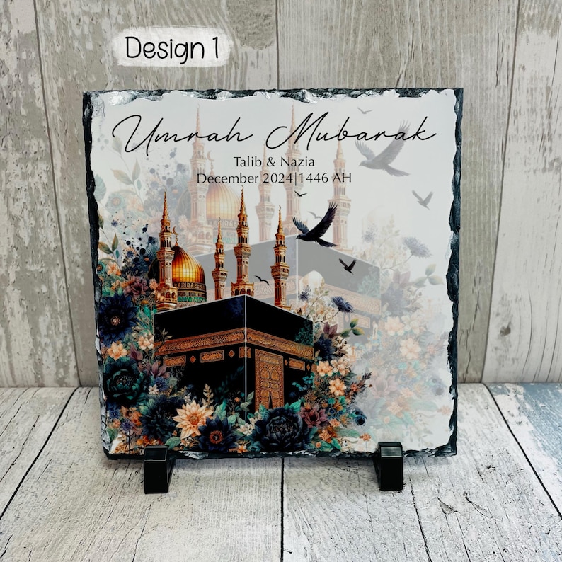 May include: A black slate plaque with a colorful image of the Kaaba in Mecca, surrounded by flowers and birds. The text "Umrah Mubarak" is written in a decorative font, along with the names "Talib & Nazia" and the date "December 2024 | 1446 AH".