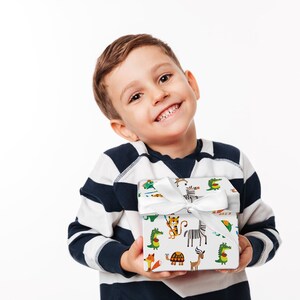 May include: A young child smiles while holding a gift box wrapped in white paper with a colorful animal print. The box is tied with a white satin ribbon. The child is wearing a navy and white striped long-sleeve shirt.