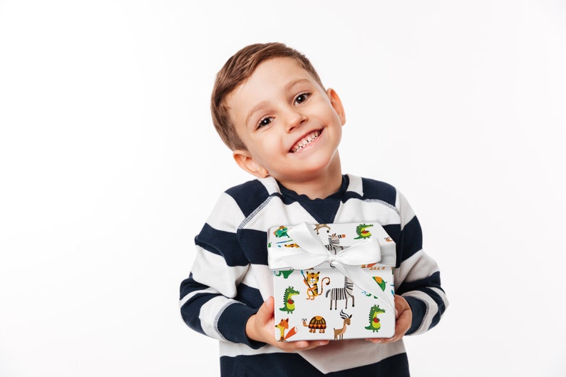 May include: A young child smiles while holding a gift box wrapped in white paper with a colorful animal print. The box is tied with a white satin ribbon. The child is wearing a navy and white striped long-sleeve shirt.