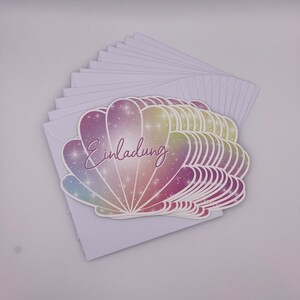 Pu&ograve; includere: Un mazzo di carte bianche vuote con un design a conchiglia colorata sulla carta superiore. La conchiglia ha un effetto glitter arcobaleno e la parola "Einladung" &egrave; scritta in corsivo viola sulla conchiglia.