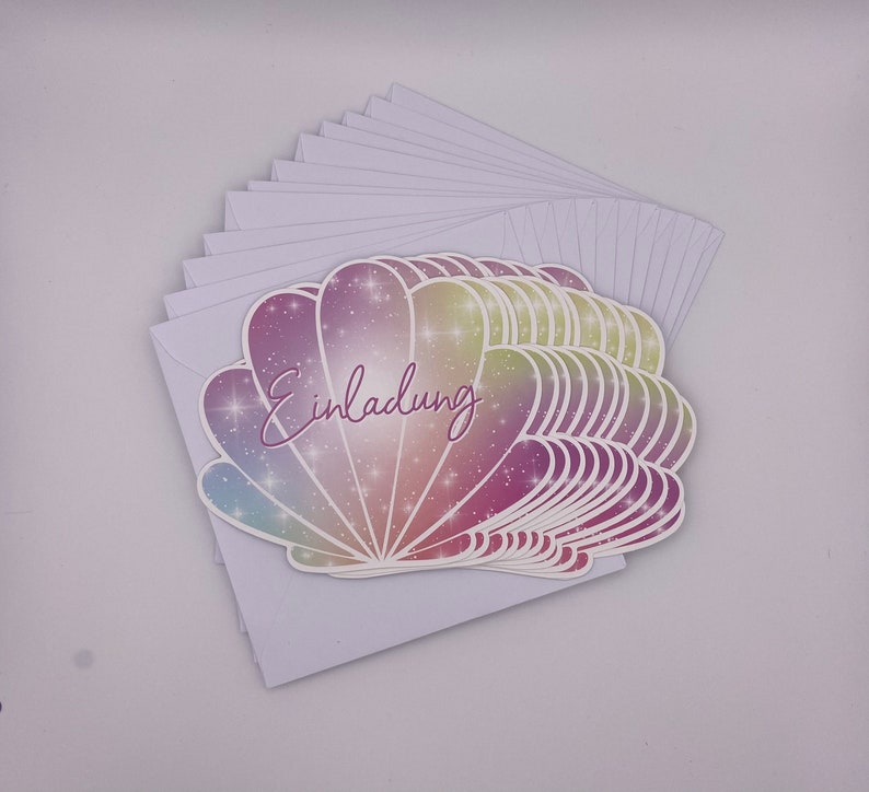 Pu&ograve; includere: Un mazzo di carte bianche vuote con un design a conchiglia colorata sulla carta superiore. La conchiglia ha un effetto glitter arcobaleno e la parola "Einladung" &egrave; scritta in corsivo viola sulla conchiglia.