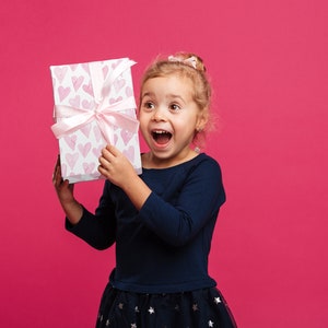 May include: A young child holds a gift box with a pink ribbon and a pattern of pink hearts. The box is white. The child is wearing a dark blue long-sleeved shirt and a dark blue skirt with star patterns, against a pink background.