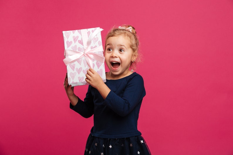 May include: A young child holds a gift box with a pink ribbon and a pattern of pink hearts. The box is white. The child is wearing a dark blue long-sleeved shirt and a dark blue skirt with star patterns, against a pink background.