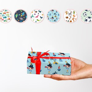 May include: A gift box wrapped in light blue paper with a pirate-themed pattern, tied with a bright red ribbon. The pattern includes cartoon pirates, ships, and palm trees. Six circular designs with various patterns are in the background.