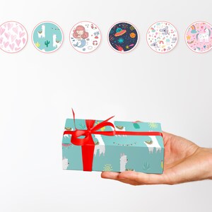 May include: A hand holding a turquoise gift box wrapped with a red ribbon. The box is decorated with a pattern of white llamas, cacti, and suns. Above the box are six circular stickers with various illustrations, including hearts, a mermaid, and a unicorn.
