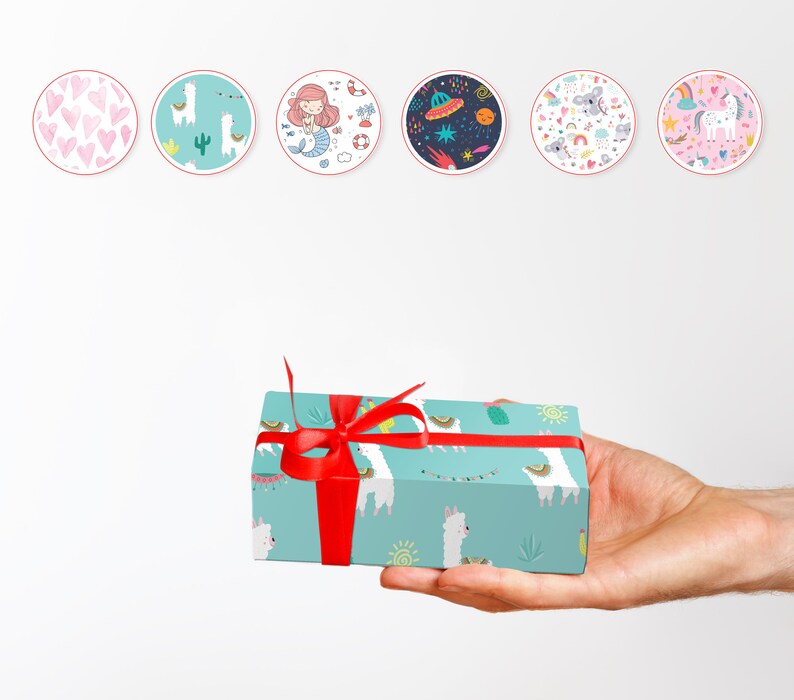 May include: A hand holding a turquoise gift box wrapped with a red ribbon. The box is decorated with a pattern of white llamas, cacti, and suns. Above the box are six circular stickers with various illustrations, including hearts, a mermaid, and a unicorn.