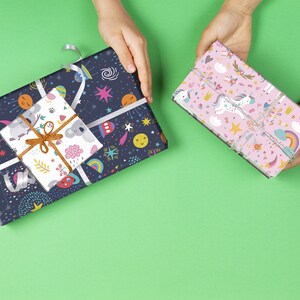 May include: Two wrapped gifts on a green background. One is wrapped in dark blue paper with a space-themed pattern, and a smaller gift on top with a koala pattern. The other gift is wrapped in pink paper with a unicorn and rainbow pattern.