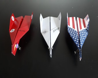 May include: Three paper airplanes, one red, one white with "USAF" printed on it, and one blue and red with stars and stripes. The airplanes are folded and have paper clips attached to them.
