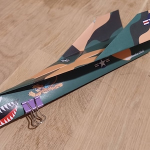 May include: A green, brown, and white paper airplane with a tiger graphic and a "Flying Tigers" logo. The airplane is held together with two purple binder clips.