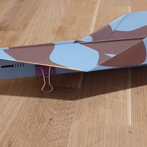 May include: A blue and brown paper airplane with a red, white, and blue flag on the tail. The airplane has a paper clip on the wing and a yellow and orange flame design on the bottom.