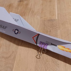 May include: A paper airplane model painted gray with USAF markings and a flame design. The plane is held together with two purple paper clips.