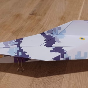 May include: A paper aeroplane with a blue and white camouflage pattern, a yellow and blue circle, and the Ukrainian coat of arms. The aeroplane is held together with a paperclip.