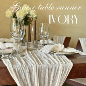 May include: A white gauze table runner with a delicate, pleated texture. The runner is decorated with a small white flower and green leaves. The text "Gauze table runner IVORY" is visible at the top of the image.
