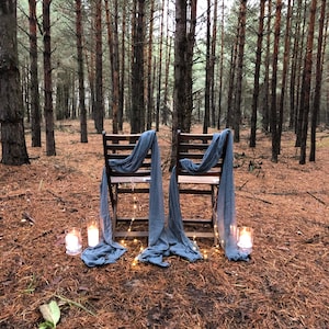 May include: Two wooden chairs with blue fabric draped over them are set up in a forest clearing. The chairs are facing each other and there are string lights and candles on the ground in front of them.