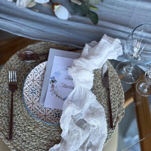 Puede incluir: Una servilleta de encaje blanco anudada en un mantel individual tejido con un plato estampado en azul y blanco. Una tarjeta blanca con el texto "The Marin" est&aacute; en el plato.