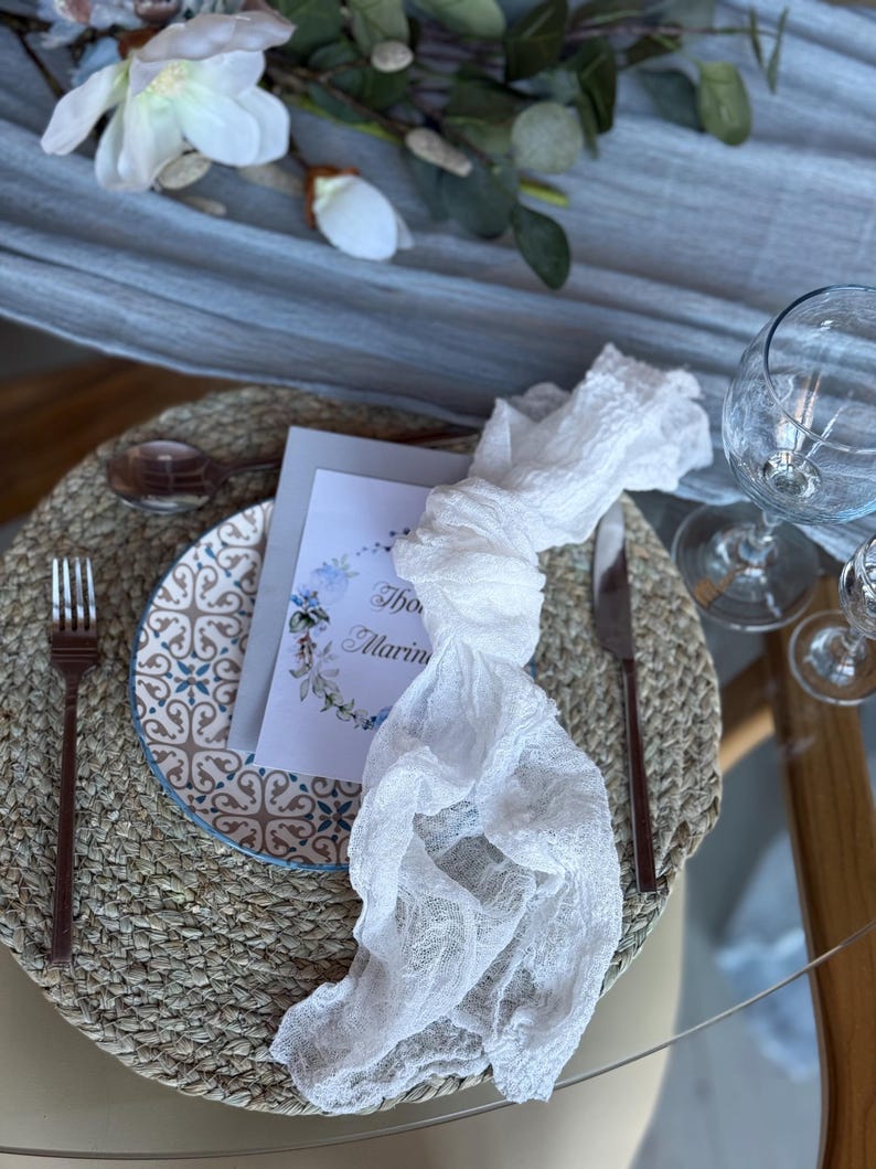 Puede incluir: Una servilleta de encaje blanco anudada en un mantel individual tejido con un plato estampado en azul y blanco. Una tarjeta blanca con el texto "The Marin" est&aacute; en el plato.