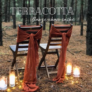 Op de afbeelding: Twee houten stoelen met terracotta gaas stoelsjerpen, vastgebonden in strikken. De stoelen staan in een bos met kaarsen en lichtslingers. De tekst "TERRACOTTA" en "Gauze chair sashes" is in het wit weergegeven.