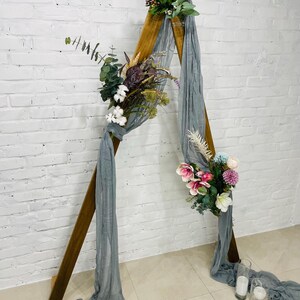 Puede incluir: Un arco de madera triangular decorado con flores blancas, rosas y violetas y vegetaci&oacute;n. Una tela transparente gris se drape sobre el arco.