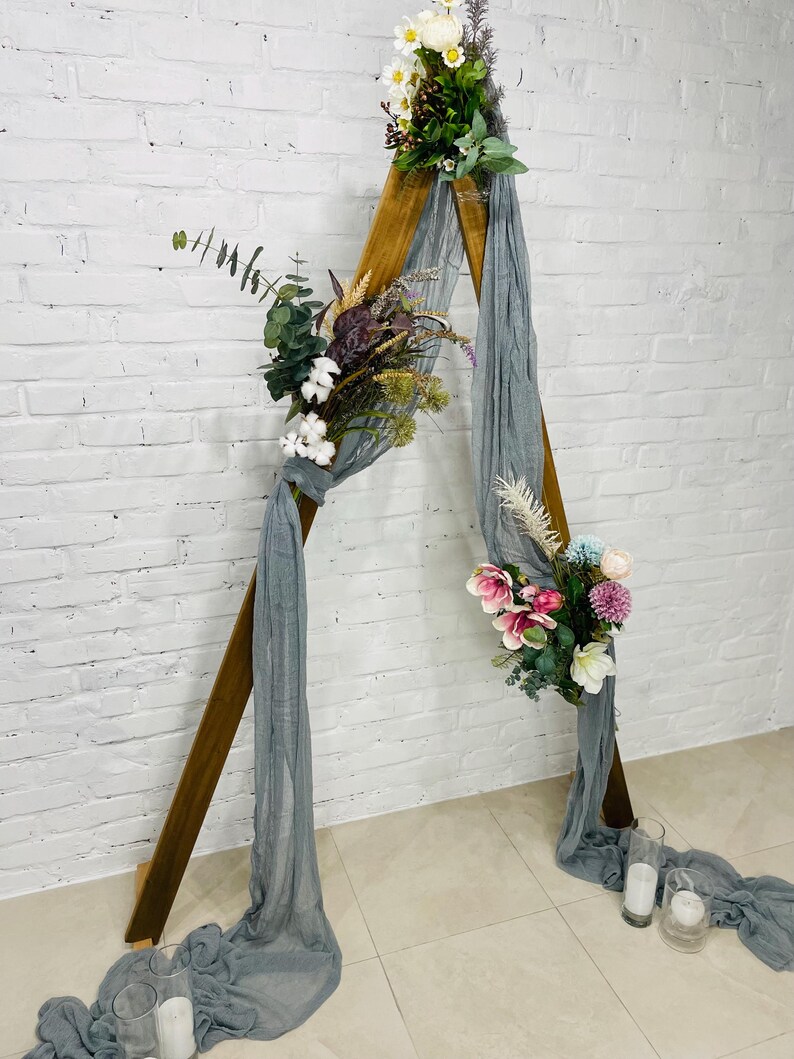 Puede incluir: Un arco de madera triangular decorado con flores blancas, rosas y violetas y vegetaci&oacute;n. Una tela transparente gris se drape sobre el arco.