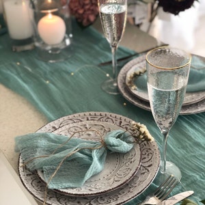 Op de afbeelding: Elegante tafelsetting met witte borden, een turquoise servet en champagneglazen met gouden randen. De borden hebben een decoratief patroon en liggen op een turquoise tafelloper. Kaarsen en bloemen.