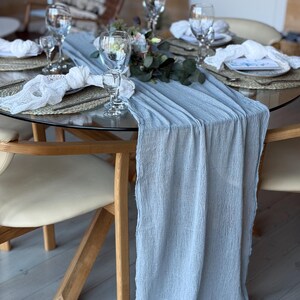 Puede incluir: Un camino de mesa azul claro con un acabado texturizado y arrugado. El camino de mesa est&aacute; colocado sobre una mesa de vidrio con una base de madera. La mesa est&aacute; puesta con platos, servilletas y vasos blancos.