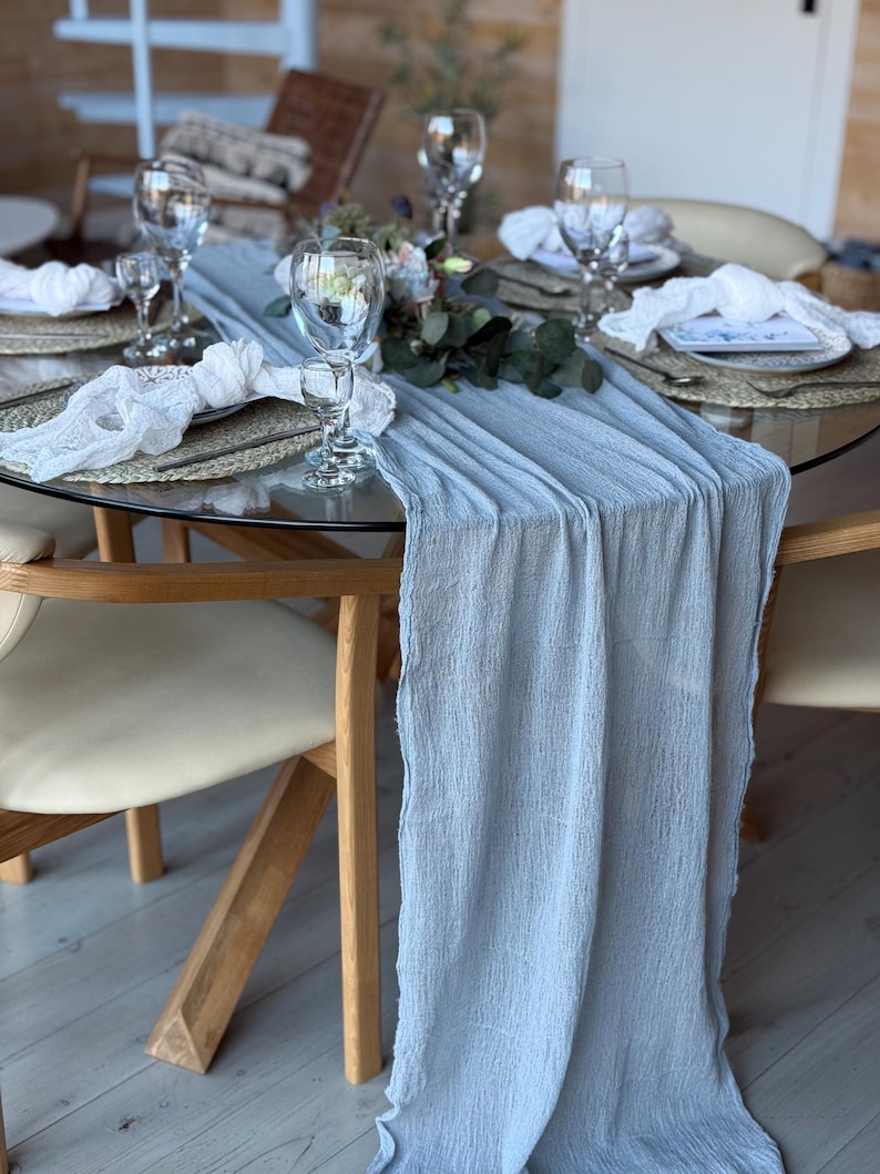 Puede incluir: Un camino de mesa azul claro con un acabado texturizado y arrugado. El camino de mesa est&aacute; colocado sobre una mesa de vidrio con una base de madera. La mesa est&aacute; puesta con platos, servilletas y vasos blancos.