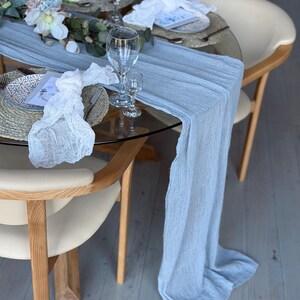 Puede incluir: Un camino de mesa azul claro con una superposici&oacute;n de encaje blanco sobre una mesa de vidrio con base de madera. La mesa est&aacute; puesta con platos, servilletas y vasos blancos. Hay flores y vegetaci&oacute;n en la mesa.