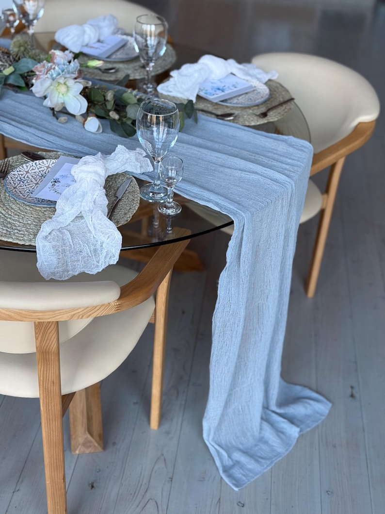 Puede incluir: Un camino de mesa azul claro con una superposici&oacute;n de encaje blanco sobre una mesa de vidrio con base de madera. La mesa est&aacute; puesta con platos, servilletas y vasos blancos. Hay flores y vegetaci&oacute;n en la mesa.